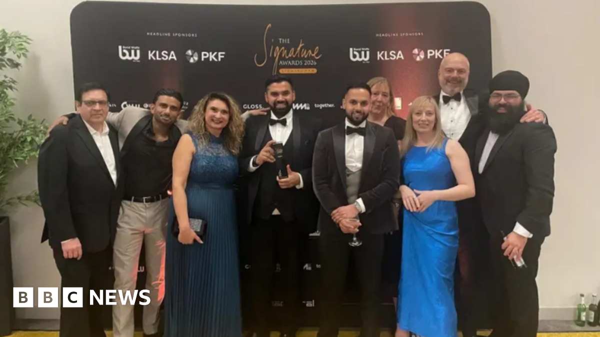 Nine men and women wearing various forms of formal evening dress pose for a picture at an awards ceremony. The man in the middle, in a dinner jacket and bow tie, is holding a black trophy. The group is standing in front of a black branded media wall featuring several sponsor names alongside the event name, The Signature Awards 2026.