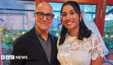Stanley Tucci stands next to a woman who is wearing a white dress and sash with the word "bride" visible. They are smiling at the camera. They are both in a restaurant with tables visible in the background.