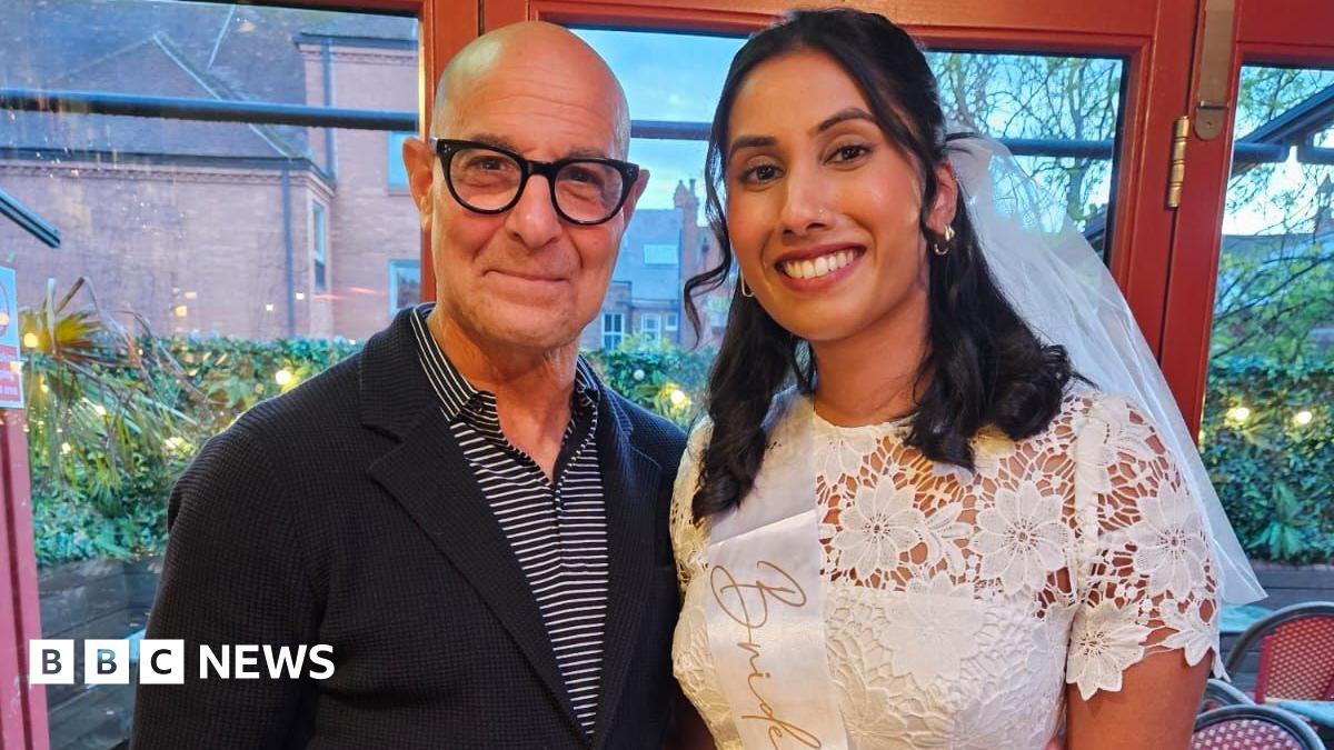 Stanley Tucci stands next to a woman who is wearing a white dress and sash with the word "bride" visible. They are smiling at the camera. They are both in a restaurant with tables visible in the background.