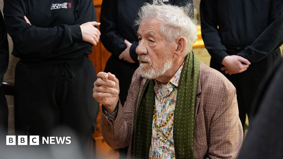 Sir Ian McKellen is an 86-year-old man wearing a tweed jacket, a green silk scarf and a flowery shirt with two long gold necklaces. He is seated and behind him are a number of people wearing black who are standing listening although only their torsos are in shot
