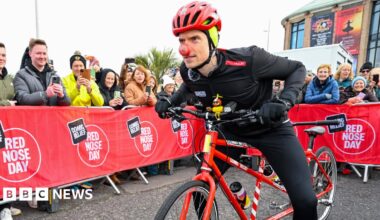 A man in black cycling gear, a red helmet and a red nose is leaning forward on a red tandem bike. There are red barriers in the street which read "Comic Relief Red Nose Day" with people stood behind them taking photographs on their phones.