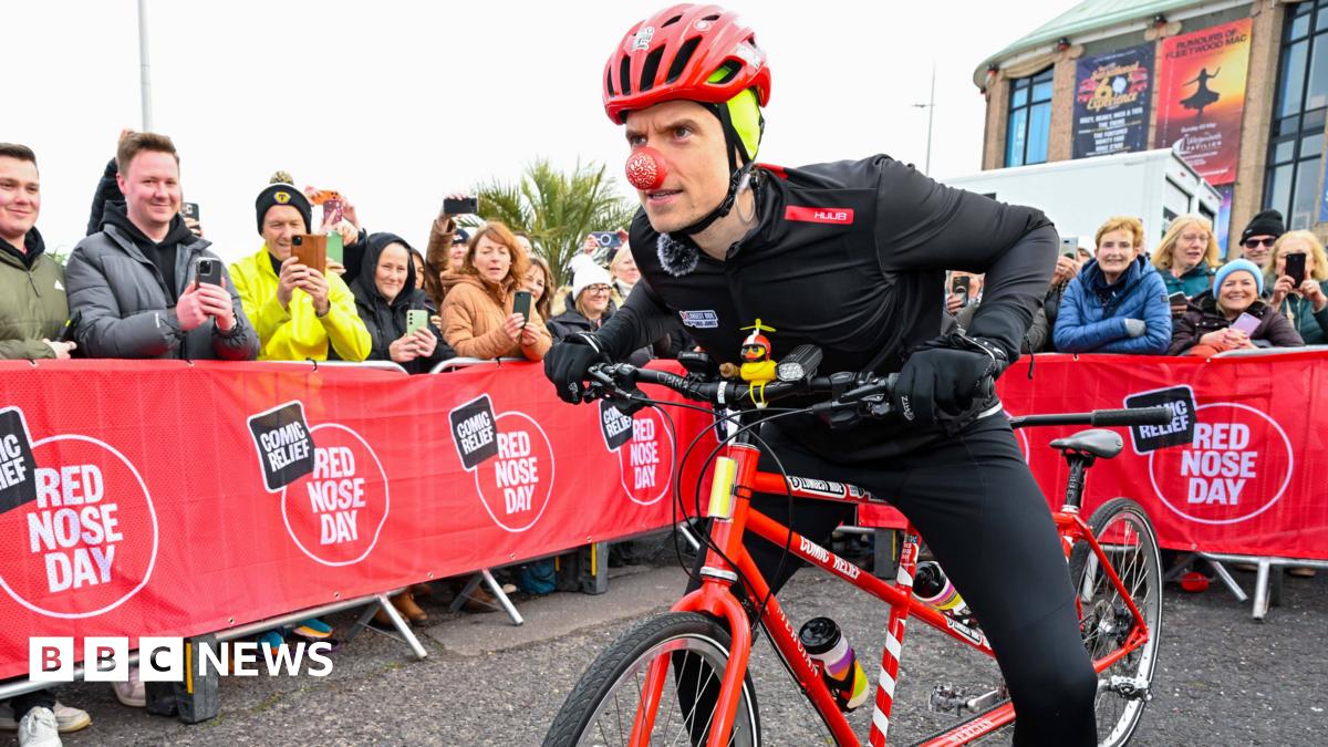 A man in black cycling gear, a red helmet and a red nose is leaning forward on a red tandem bike. There are red barriers in the street which read "Comic Relief Red Nose Day" with people stood behind them taking photographs on their phones.