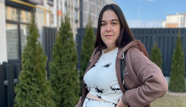 A pregnant woman stands in a garden with her hand on her belly. She has long, straight, dark hair and is wearing a matching top and trousers which are white with some patches of black. She also has a light brown hoodie on.