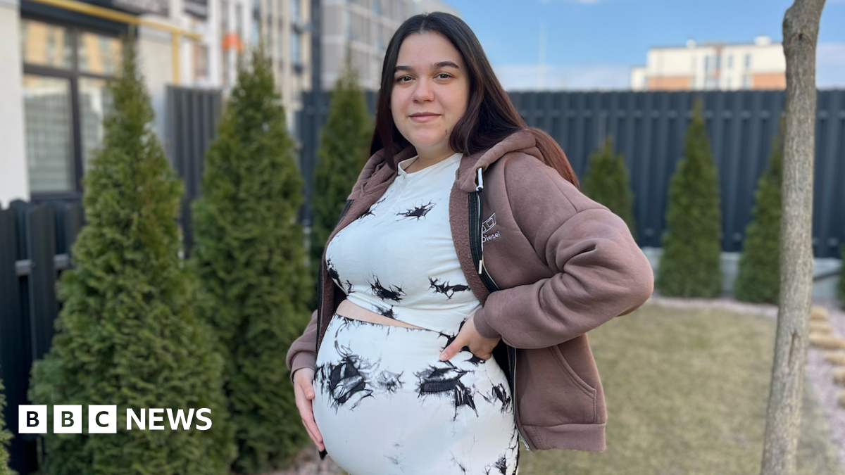 A pregnant woman stands in a garden with her hand on her belly. She has long, straight, dark hair and is wearing a matching top and trousers which are white with some patches of black. She also has a light brown hoodie on.