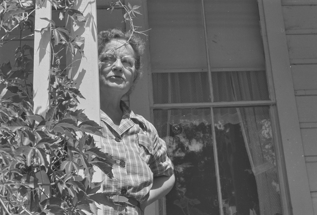 An older woman wearing glasses and a plaid shirt stands by a vine-covered porch post, looking upward in bright sunlight.