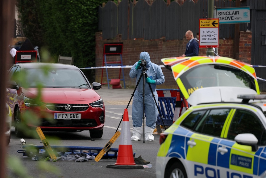 A crime scene technician stands away from some debris on the ground and photographs it. He's wearing a full body suit.