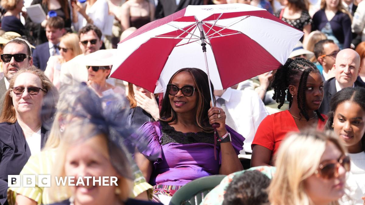 Crowd of people sitting in the Sun at event. Two people in centre holding an umbrella to shield themselves from the Sun