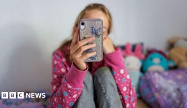 A anonymous young girl looking at her phone on her bed in a room with brightly coloured cuddly toys. She is wearing a pink flowery jumper and jeans, we cannot see her face.