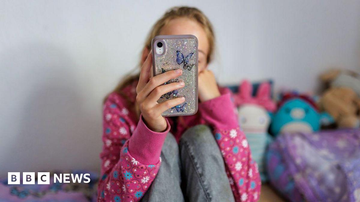A anonymous young girl looking at her phone on her bed in a room with brightly coloured cuddly toys. She is wearing a pink flowery jumper and jeans, we cannot see her face.