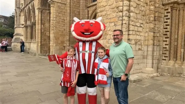 The club's imp mascot poses with the two children who wear scarves and football shirts and flags.  Their father stands at the side of them.
