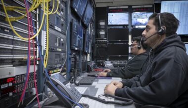 Two men wearing headsets sit in front of a wall of screens and instrumentation.