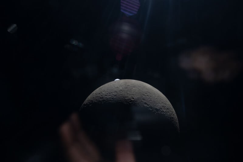 A dark, shadowed view of the Moon’s curved surface with a small, bright Earth visible rising above the horizon in the background, set against the blackness of space.