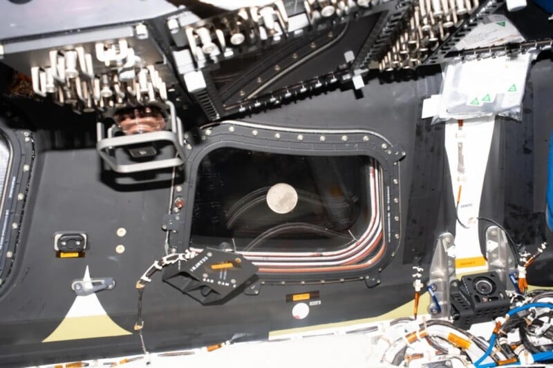 A view of the Moon through a window from inside a spacecraft cabin, surrounded by panels, instruments, and cables. The Moon appears centered and bright against the dark window.