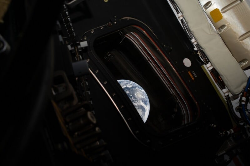 View of Earth seen through a rectangular window from inside a spacecraft, with surrounding equipment visible in the dimly lit interior.