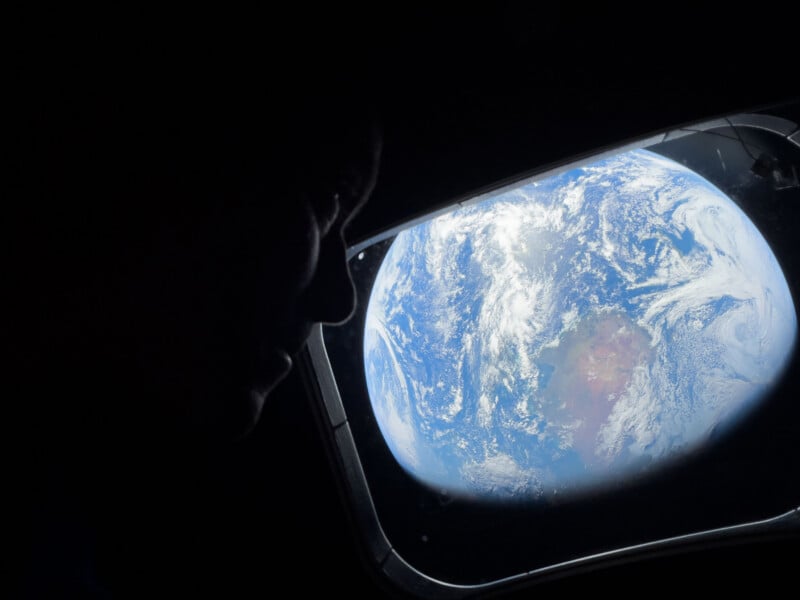 A person’s silhouette is seen looking out of a spacecraft window at Earth, which appears bright and full of clouds and blue oceans against the darkness of space.