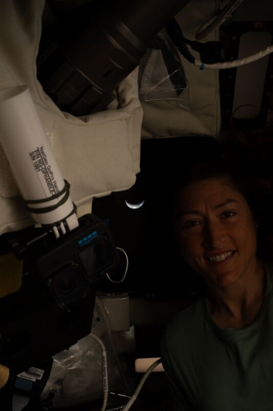 A person smiles inside a dimly lit spacecraft with equipment visible. Through a window behind them, a crescent Earth is seen against the darkness of space.