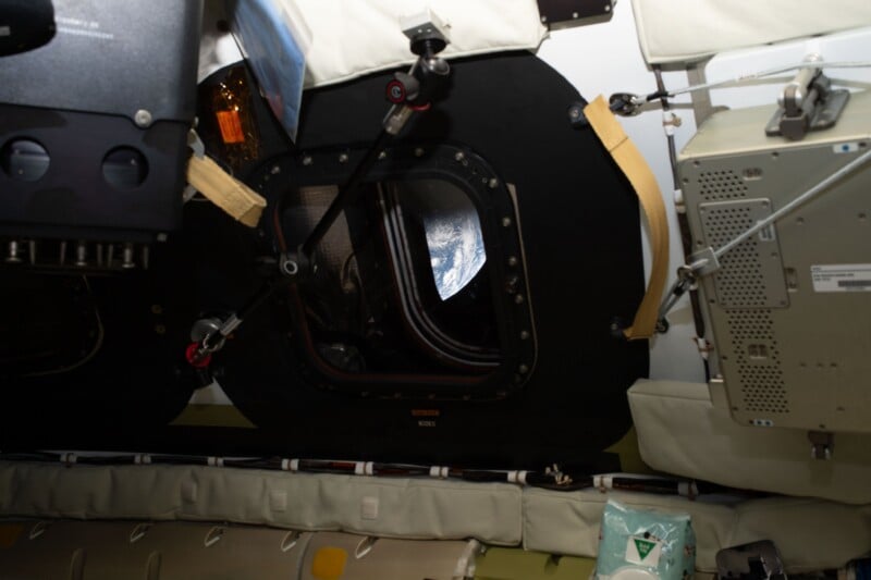 View of Earth through a small round window inside a spacecraft, surrounded by complex equipment, panels, and cushions. The partially visible planet is framed by the spacecraft's dark interior.