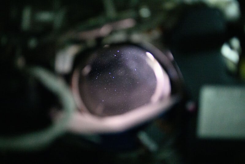A view of a starry night sky seen through a round window, possibly from a spacecraft or space station, with blurred edges and equipment visible around the window frame.
