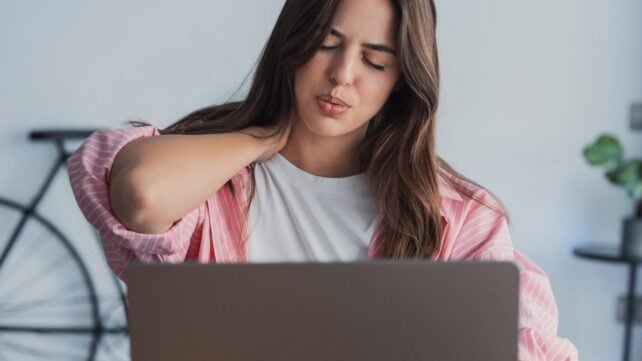 Woman sitting at a laptop massages the back of her neck in discomfort