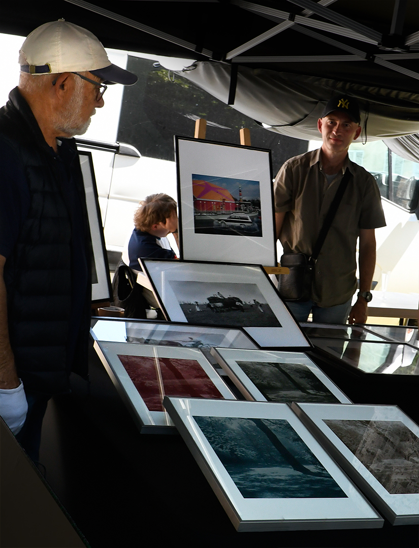 A visitor in a baseball cap examines framed photographic prints at an artist's booth, with colour and black-and-white works displayed across a glass-topped table.