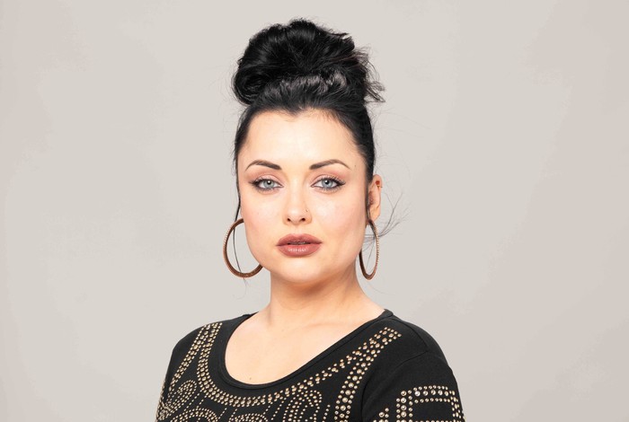 Eastenders Whitney exit Shona McGarty poses as Whitney Dean in an EastEnders promo image wearing a black pearled top and hoop earrings.
