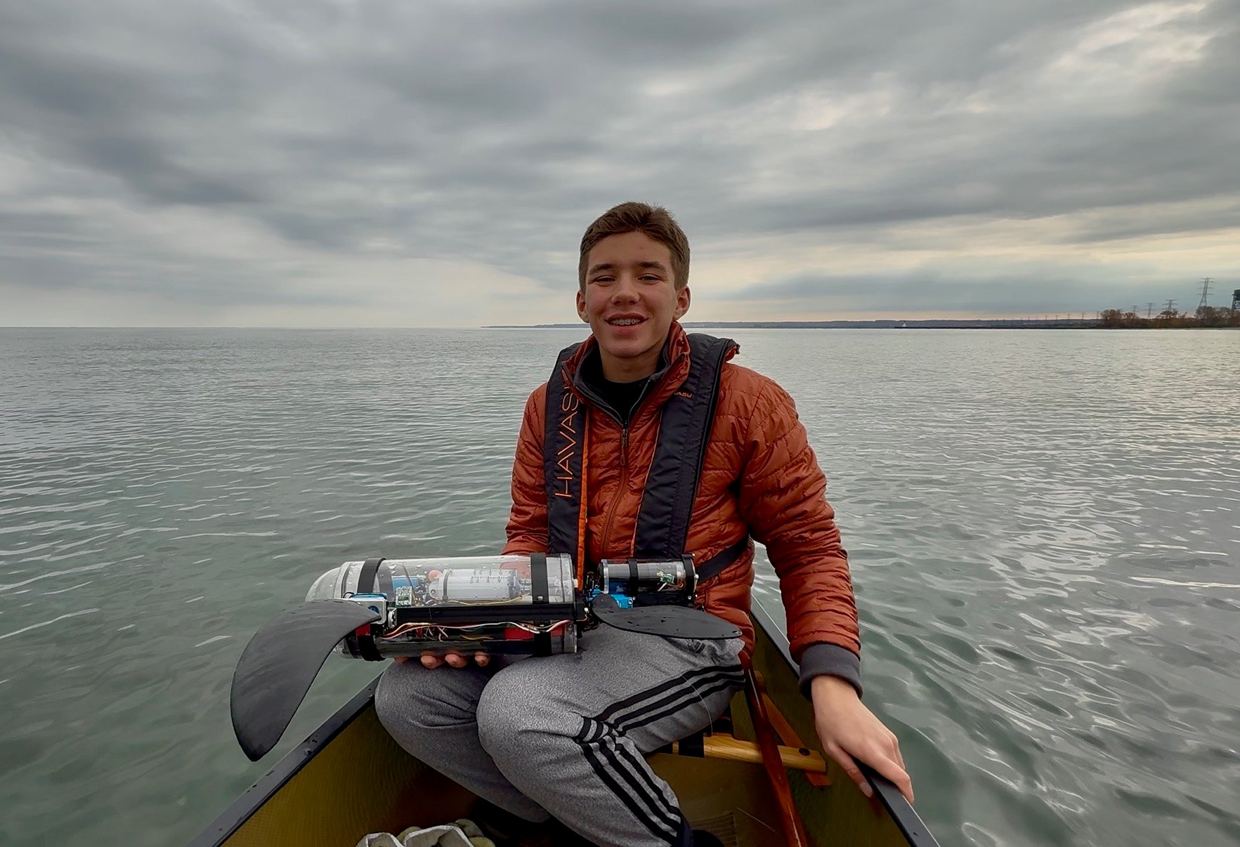 A boy in an orange puffer jacket holds a bionic turtle while sitting in a canoe in a lake.