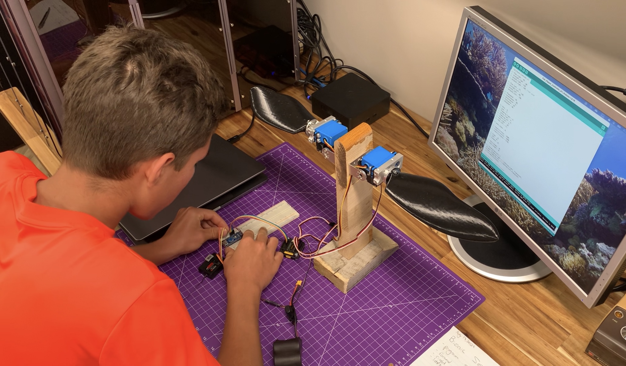 Boy in orange T-shirt works on robotic flippers in front of a computer.