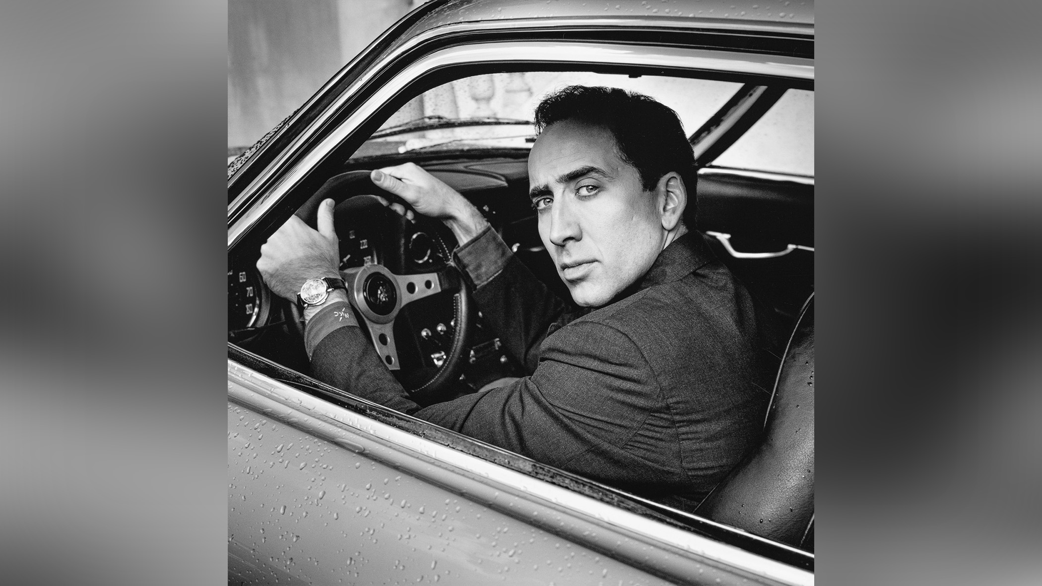 A man sat in the driver's seat of a car looks out the window toward the camera.