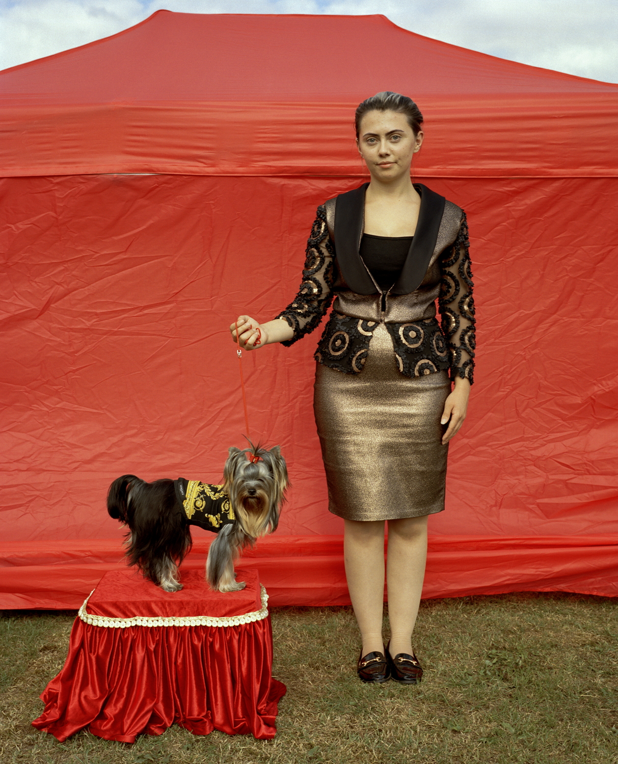 A young woman in a gold lamé skirt and ornate beaded jacket holds the lead of a Yorkshire Terrier posed on a red satin plinth, both standing before a vivid red pop-up tent.