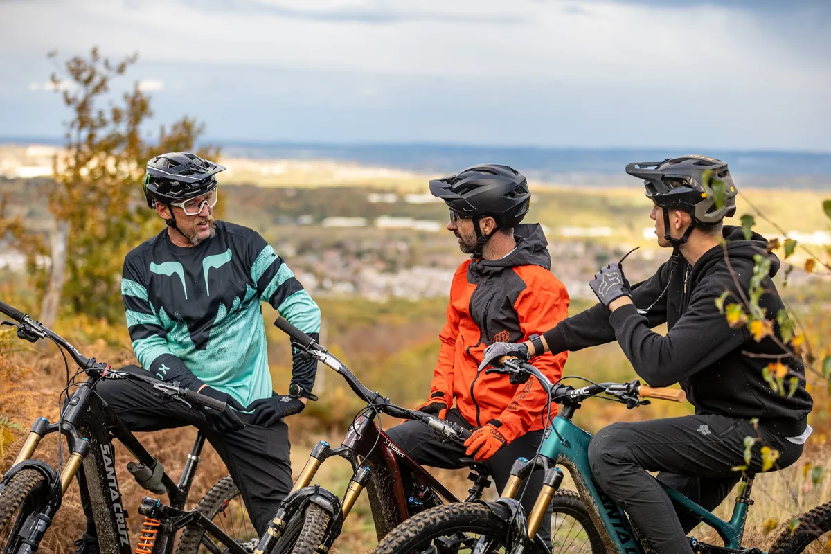 Three mountain bikers wearing helmets pause on a hillside trail with a town and coastline visible in the background.