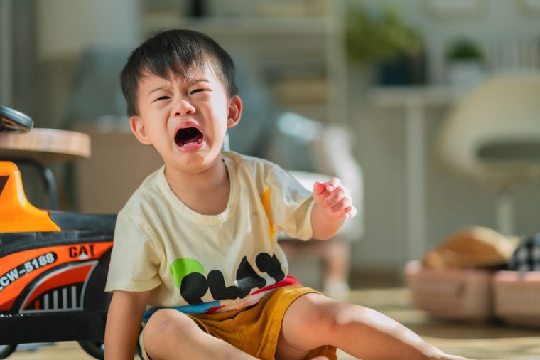 Asian small toddler child is riding a toy car when he falls on the floor, leading to cries and shouts. The unhappy boy, playing with his toy car in the living room daylight, sheds tears - stock photo