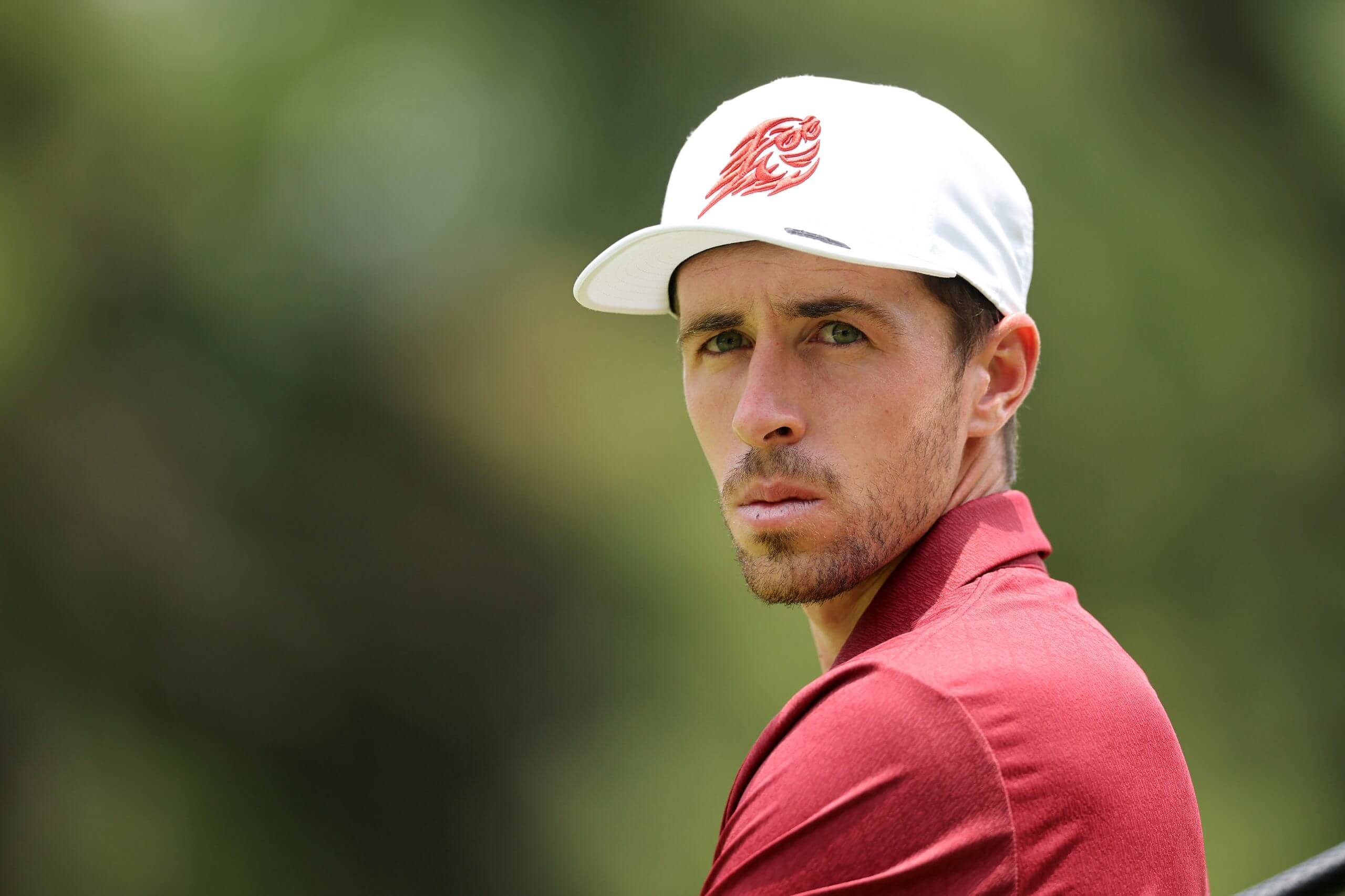 Close-up of David Puig looking to his left in a red shirt, white hat and with trees blurred in the background.