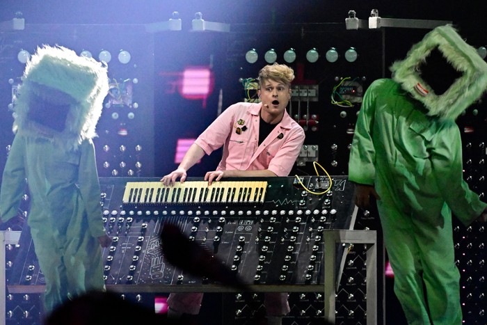 A blone man wearing pink boiler suit pulling a shocked expression behind a DJ deck.