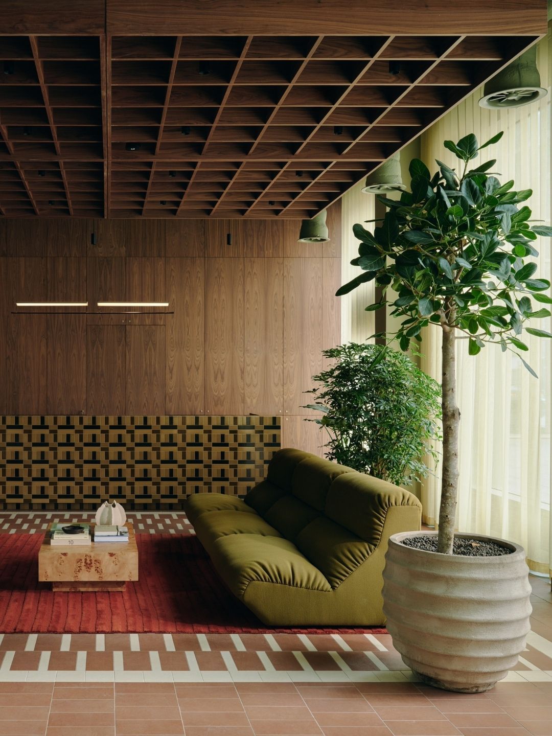 Image of an olive green sofa in a mid-century modern-inspired space with wooden walls and a red rug and lots of plants.