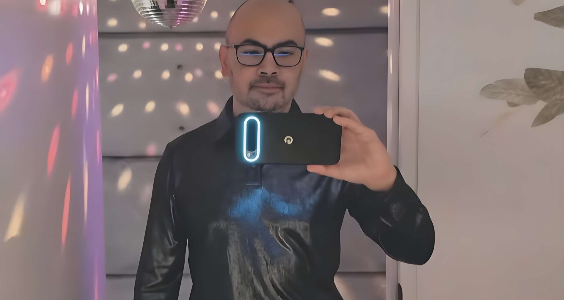 A person wearing a black shirt takes a selfie in a room with a disco ball and colorful light reflections, holding a smartphone with a glowing camera module.