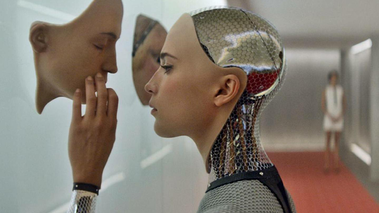 Still from the movie " Ex Machina." Close up of an android with a human face is touching a human face hanging on a wall.
