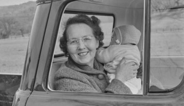 A smiling woman wearing glasses sits in the open window of a car, holding up a bundled infant against a wintry rural backdrop.