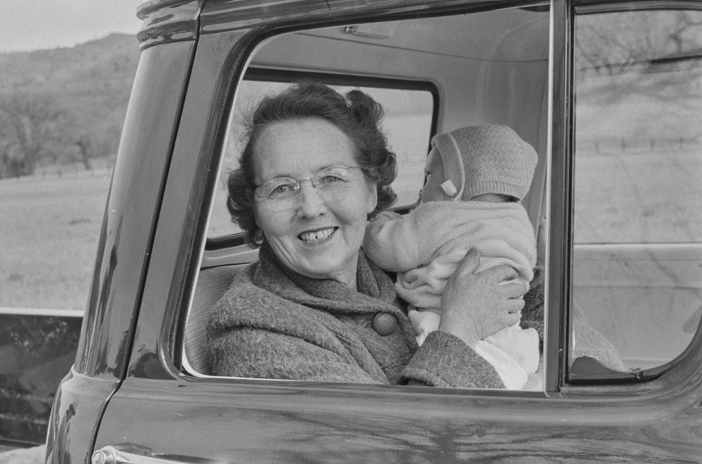A smiling woman wearing glasses sits in the open window of a car, holding up a bundled infant against a wintry rural backdrop.