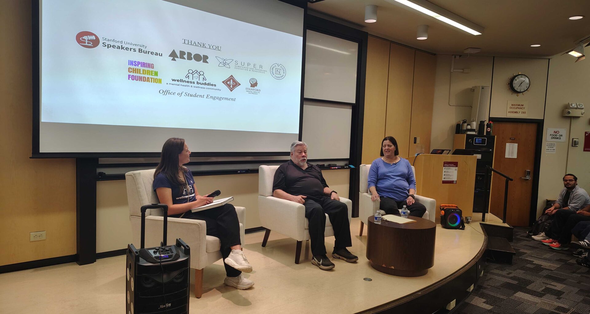 Panelists sit in armchairs on a wood-paneled stage.