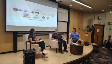 Panelists sit in armchairs on a wood-paneled stage.