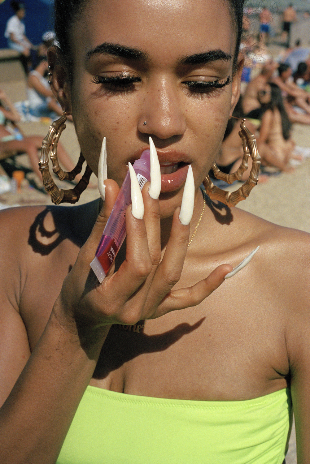 A close-up portrait of a young woman in a neon yellow bandeau top applying lip gloss with dramatically long white stiletto nails, large gold bamboo hoop earrings framing her face on a crowded beach.