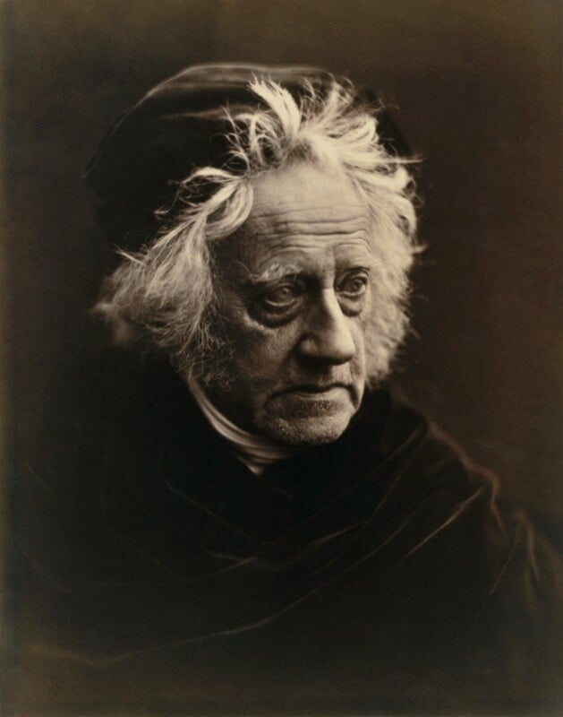 Sepia-toned portrait of an elderly man with wild white hair, wearing a dark cloak and cap, gazing thoughtfully to the side against a plain background.