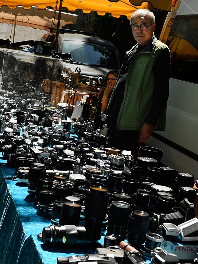 A vendor stands behind a long table blanketed in vintage SLR cameras, lenses and accessories, including a Pentacon, laid out on a blue cloth.
