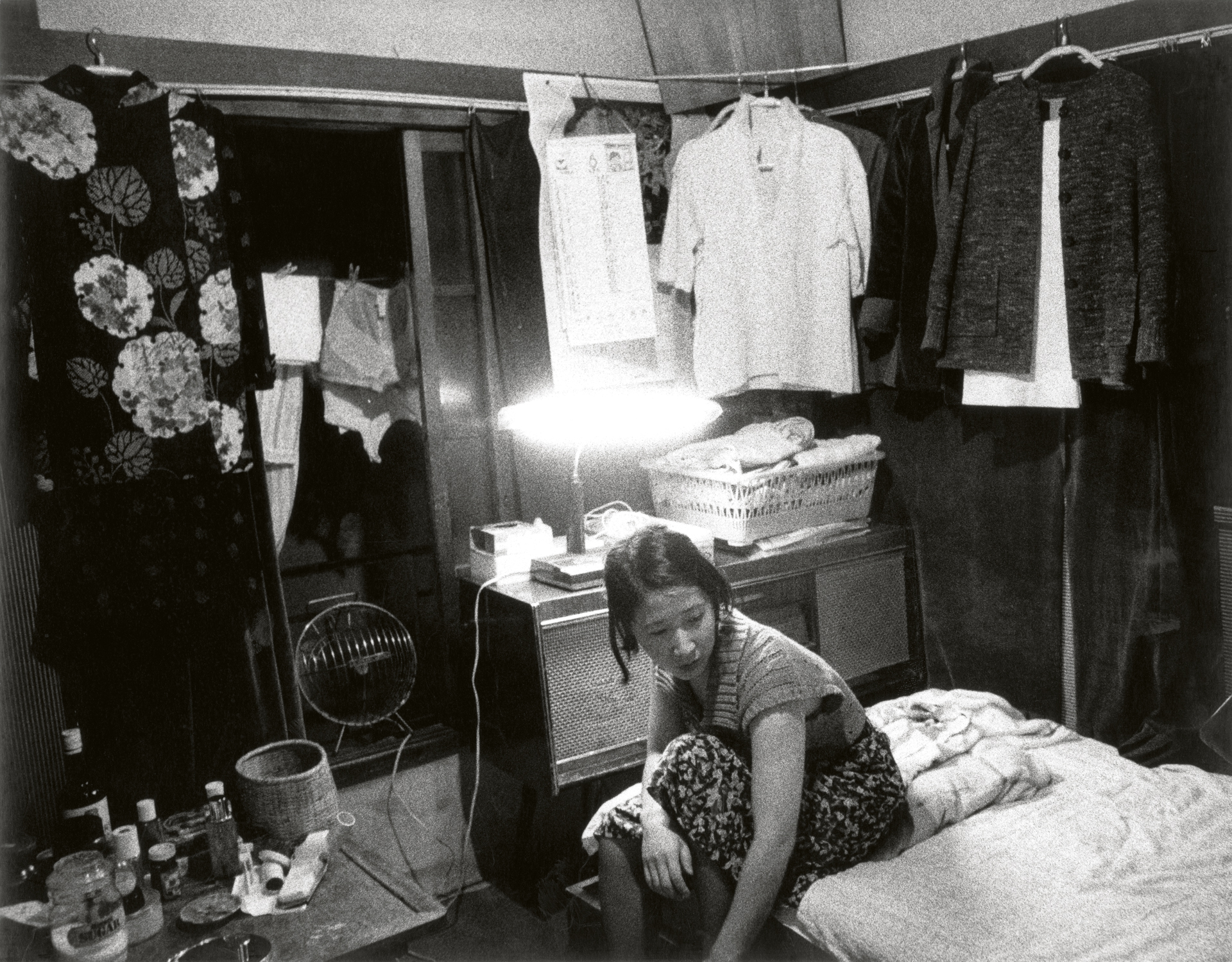 Black and white interior photograph of a woman sitting on a bed in a cramped apartment with laundry hanging on a clothesline above her.