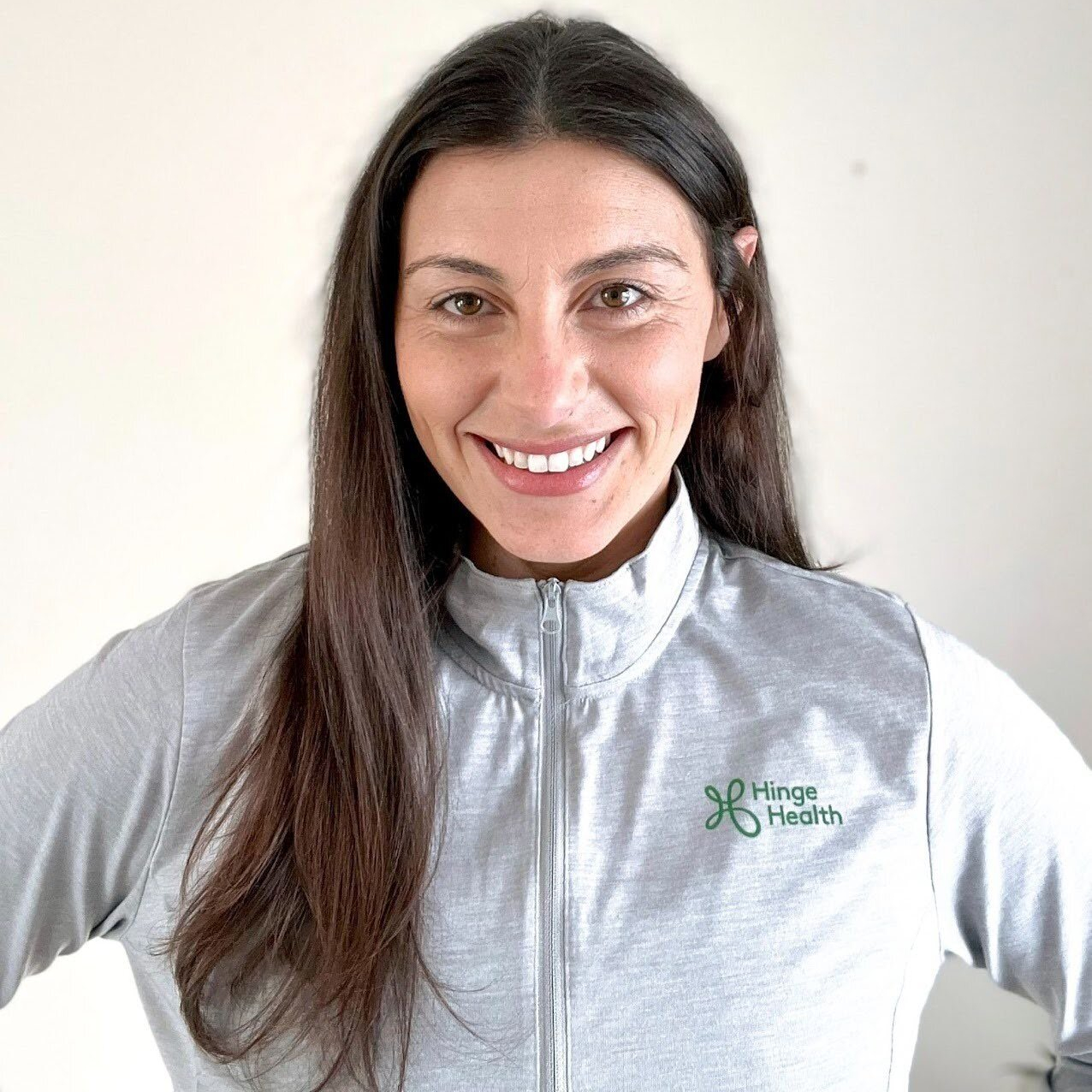 Woman with long brown hair and a zip-up gray long-sleeved top with the Hinge Health logo on it smiles at camera