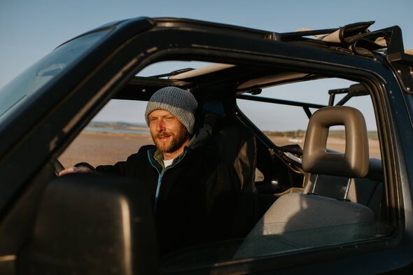 A smiling man wearing casual clothing, including a beanie hat, drives an off road vehicle on a beach on a sunny evening. He look A smiling man wearing casual clothing, including a beanie hat, drives an off road vehicle on a beach on a sunny evening. He look