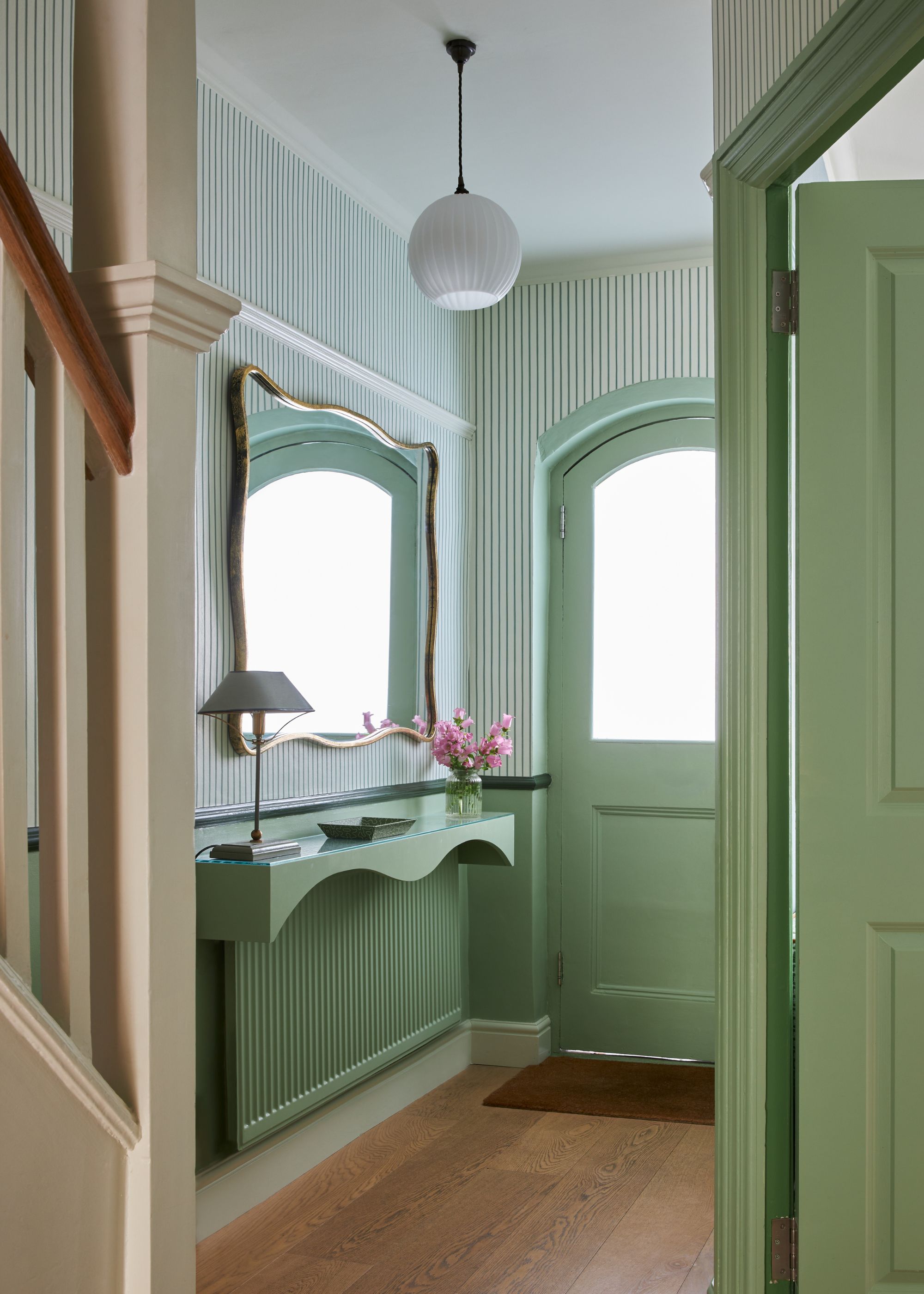 An entryway with wooden flooring, a light green arched front door, a green shelf above a green radiator with a mirror, and striped light green and white wallpaper on the upper walls.