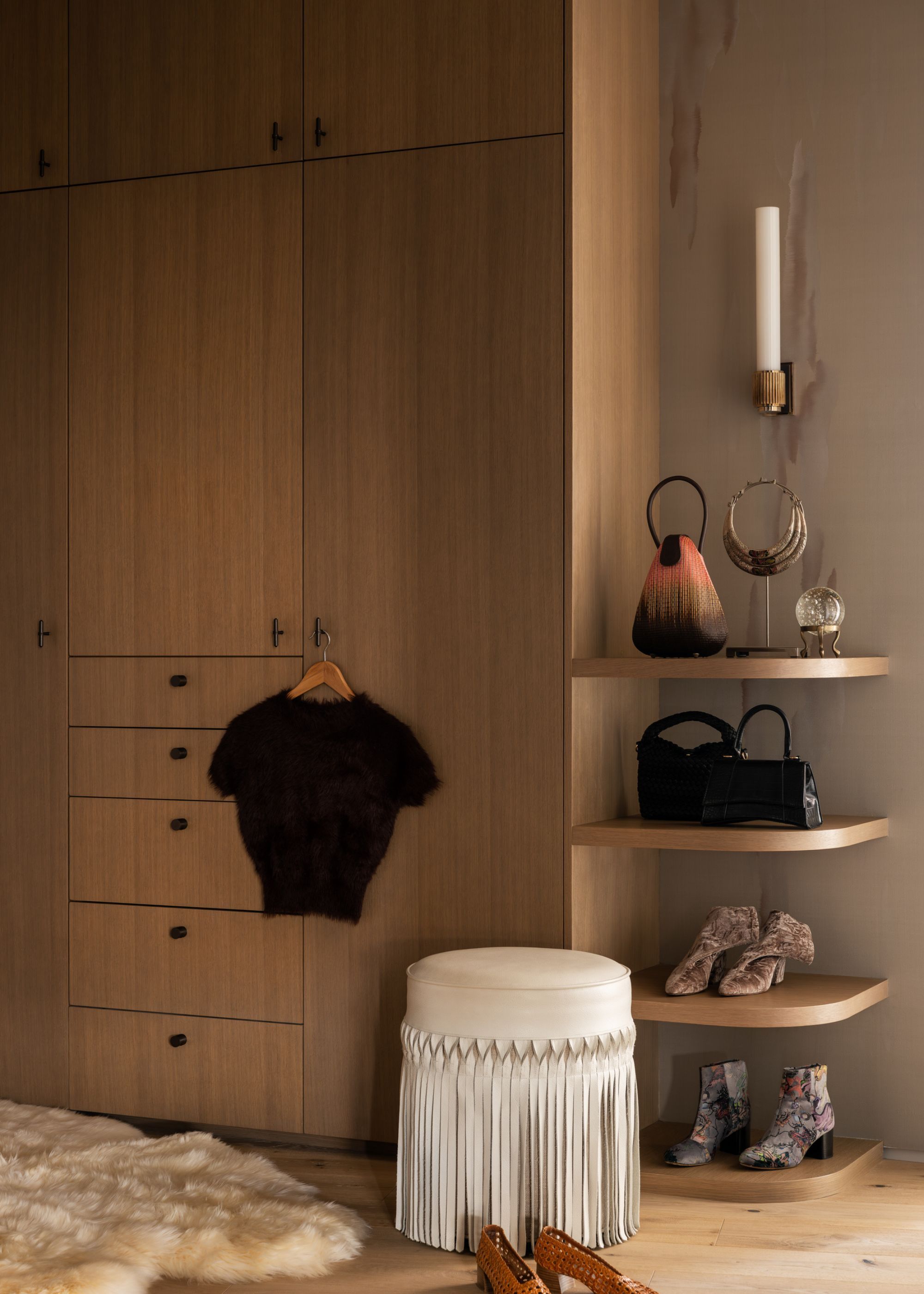 A dressing are with light-tones timber built-in closets and decorative shelving to display shoes and bags with a white fringe-trimmed stool in front of it next to a sheepskin rug