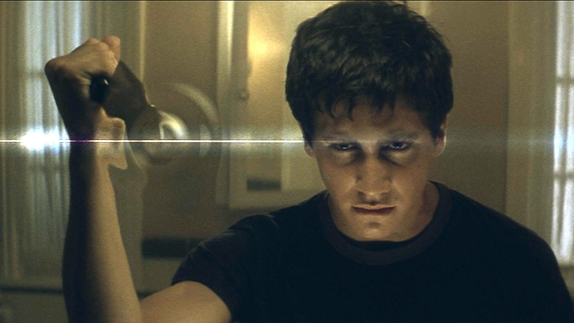 A young man with dark circles around his eyes and dark shaggy hair is holding an object in a mirror