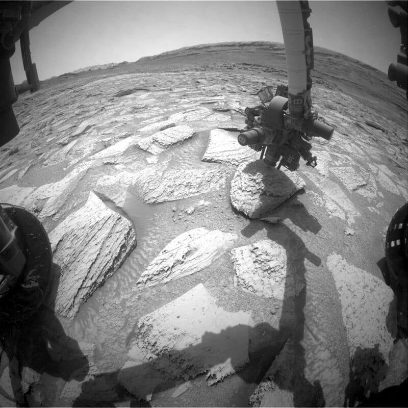 Black and white image of the Mars Curiosity rover’s robotic arm extended over cracked, flat rock slabs on the Martian surface, with part of the rover’s wheels visible in the foreground. The landscape is barren and dusty.